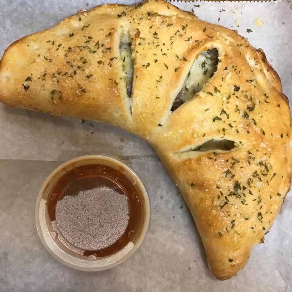 Golden-brown calzone with visible cheese and meat filling, garnished with herbs, next to a cup of marinara sauce.
